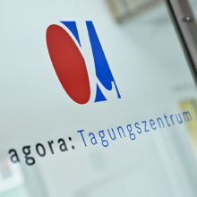Eine Glastür mit dem Logo und dem Text agora: Tagungszentrum auf Deutsch. Das Logo besteht aus einem roten Oval und einer blauen abstrakten Form über dem Text. Die Metallrahmen der Tür sind teilweise sichtbar.