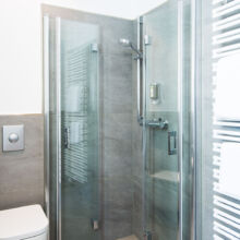 Ein modernes Badezimmer mit einer Glasduschkabine, einer grau gefliesten Wand, einer wandmontierten Toilette, einem vertikalen Handtuchwärmer mit gefalteten weißen Handtüchern und einem verchromten Duschkopf.