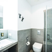 Modernes Badezimmer mit rechteckigem Waschbecken, Glasduschabtrennung, wandmontierter Toilette und minimalistischem Dekor in Grau und Weiß. Zwei verspielte schwarze Wandhaken in Form von Kletterfiguren befinden sich in der Nähe der Dusche.