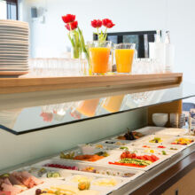 Ein Frühstücksbuffet mit Aufschnitt, Käse, Obst, Gemüse und Gebäck. Darüber stapeln sich Teller, Gläser, rote Tulpen in Vasen und zwei Krüge mit Orangensaft. Milchkartons stehen seitlich auf dem Tresen.