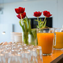 Nahaufnahme eines Holztisches, auf dem Krüge mit Orangensaft, Reihen von leeren Gläsern, gestapelte weiße Tassen und eine Vase mit roten Tulpen stehen. Der Hintergrund erscheint leicht unscharf.
