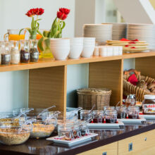 A breakfast buffet with jars of cereals, bowls of jams, Nutella, stacked plates, glasses, white bowls, baskets of bread, and two vases with red tulips on a wooden counter.