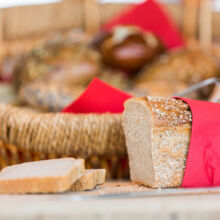 Ein in rotes Papier eingewickelter Laib Haferbrot liegt auf einem Holzbrett, in das zwei Scheiben geschnitten sind. Im Hintergrund steht ein Weidenkorb mit verschiedenen Brotsorten, die ebenfalls in rotes Papier eingewickelt sind.
