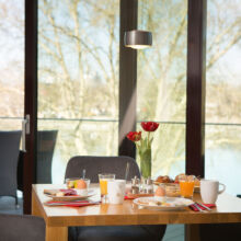Ein für das Frühstück gedeckter Holztisch mit Tellern mit Speisen, Saft, Kaffee, einem weich gekochten Ei und roten Tulpen in einer Vase. Hinter großen Fenstern sieht man Bäume und einen Balkon mit Stühlen im Freien.