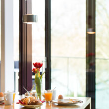 Ein heller Frühstückstisch, gedeckt mit Croissants, Eiern, Saft, Kaffee und einer Vase mit roten Tulpen, neben großen Glasfenstern mit Blick auf eine verschwommene Außenansicht.