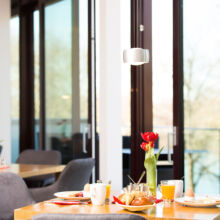 Ein heller, moderner Essbereich mit gedeckten Tischen für das Frühstück mit Croissants, Eiern, Saft, Kaffee und roten Tulpen in der Nähe von großen Fenstern mit Blick auf einen malerischen Außenbereich.