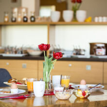 Ein Frühstückstisch, gedeckt mit Kaffee, Saft, Eiern, Brot, Müsli und Obst, mit zwei roten Tulpen in einer Vase. Im Hintergrund ist ein Buffet mit verschiedenen Lebensmitteln auf einer Theke zu sehen.
