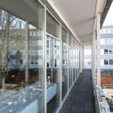 Ein langer, schmaler Balkon mit Glaswänden führt an einem modernen Gebäude entlang. In dem Glas spiegeln sich Bäume und nahe gelegene Gebäude. Auf dem Boden darunter sind mehrere Fahrräder geparkt.