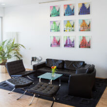 Ein modernes Wohnzimmer mit schwarzen Ledersesseln und -sofas, einem gläsernen Couchtisch mit einer Vase mit Tulpen, einem großen schwarzen Teppich, einer Topfpflanze und bunten geometrischen Kunstwerken, die in einem Raster an der Wand angeordnet sind.