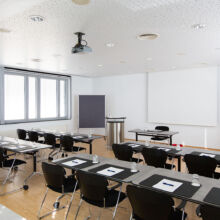 Ein moderner, gut beleuchteter Konferenzraum mit schwarzen Stuhlreihen und Tischen, die mit Notizblöcken, Stiften und Gläsern ausgestattet sind. Es gibt ein Podium, einen Projektor, eine große weiße Leinwand und Fenster an einer Wand.