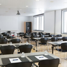 Ein modernes, gut beleuchtetes Klassenzimmer mit Reihen von Tischen und schwarzen Stühlen, jeder Tisch mit einem Notizblock, einem Stift und einem Glas Wasser ausgestattet. Auf einer Seite befinden sich große Fenster und an der Decke ist ein Projektor angebracht.