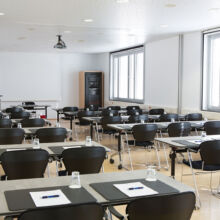 Ein modernes, gut beleuchtetes Klassenzimmer mit Reihen von Tischen und schwarzen Stühlen, jeder Tisch mit einem Notizblock, einem Stift und einem Glas Wasser ausgestattet. Auf einer Seite befinden sich große Fenster und an der Decke ist ein Projektor angebracht.