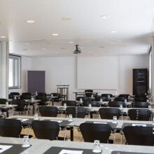 Ein moderner, gut beleuchteter Konferenzraum mit Reihen schwarzer Stühle und Tischen, die mit Notizblöcken, Stiften und Wassergläsern bestückt sind und auf eine leere Projektionsfläche im vorderen Teil des Raums blicken.