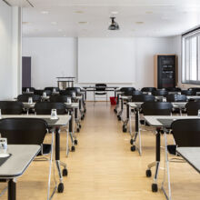 Ein modernes, leeres Klassenzimmer mit Reihen schwarzer Stühle und Tische, jeder mit einem Notizblock, einem Stift und einem Glas Wasser ausgestattet. Große Fenster auf der rechten Seite lassen natürliches Licht herein, und an der Decke ist ein Projektor angebracht.