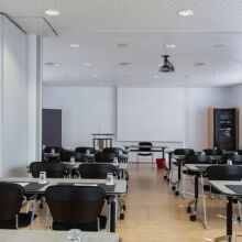 Ein helles, modernes Klassenzimmer mit Reihen von schwarzen Stühlen und Tischen, Gläsern auf jedem Tisch, einem Podium an der Stirnseite und großen Fenstern, die Tageslicht hereinlassen.