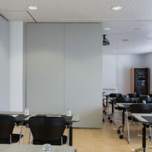 Ein modernes Klassenzimmer mit schwarzen Stühlen und in Reihen angeordneten Tischen, Glasschalen auf jedem Tisch, einer Trennwand, die den Raum unterteilt, und großen Fenstern auf der rechten Seite, die natürliches Licht hereinlassen.
