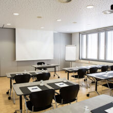 Ein moderner Konferenzraum mit Reihen von Tischen und Stühlen vor einer Projektionsfläche, mit Notizblöcken, Stiften und Wassergläsern auf jedem Tisch. Große Fenster lassen von der rechten Seite natürliches Licht herein.