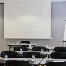 Ein Konferenzraum mit Reihen von Tischen und Stühlen, die jeweils mit einem Notizblock, einem Stift und einem umgedrehten Glas ausgestattet sind. An der Stirnseite befinden sich eine leere Projektionsfläche und ein Flipchart mit leerem Papier. Der Raum scheint bereit für eine Besprechung oder Präsentation.