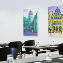 Ein modernes Klassenzimmer mit schwarzen Stühlen und Tischen, jeder Tisch mit einem Glas und einem Notizblock ausgestattet. An der weißen Wand im Hintergrund hängen zwei bunte Gemälde von Gebäuden.