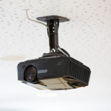 Ein schwarzer, an der Decke montierter Projektor ist mit einer Metallhalterung an einer weißen, perforierten Decke befestigt. Ein weißes Stromkabel führt vom Projektor in die Decke.