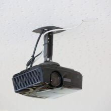 Ein schwarzer, an der Decke montierter Projektor ist an einer weißen, durchlöcherten Decke befestigt und mit sichtbaren Kabeln verbunden.