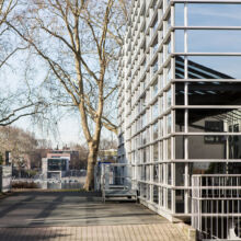 Ein modernes Glasgebäude mit einer Metallgitterfassade steht an einem sonnigen Tag neben einem gepflasterten Weg und blattlosen Bäumen, im Hintergrund sind Wasser und weitere Gebäude zu sehen.