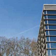 Rechts ist ein modernes Gebäude mit großen Glasfenstern zu sehen, in denen sich der blaue Himmel spiegelt. Auf der linken Seite stehen blattlose Bäume, und über dem Himmel kreuzt ein schwacher Kondensstreifen den Himmel.