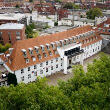 Außenansicht des Hotel agora: am Aasee. Ein großes weißes Gebäude mit rotem Ziegeldach und mehreren Dachgauben, umgeben von Bäumen und anderen Gebäuden in einer städtischen Umgebung.