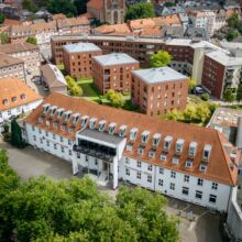 Außenansicht des Hotel agora: am Aasee. Luftaufnahme eines weißen Gebäudes mit rotem Ziegeldach und Reihen von Dachgauben, umgeben von modernen Backsteinwohnungen und Bäumen in einem städtischen Viertel.