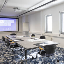 Ein moderner Konferenzraum mit einem langen Tisch, acht Stühlen, Notizblöcken und Stiften, die fein säuberlich angeordnet sind. An der Vorderseite befinden sich eine Projektionsfläche, ein Flipchart und zwei große Fenster, durch die natürliches Licht einfällt.