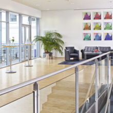 Moderne Lobby mit Glaswänden, kleinen runden Tischen mit Wasserflaschen, einer schwarzen Sofagarnitur, einem Sessel, einer Topfpflanze und bunten Kunstwerken an der weißen Wand. Sonnenlicht erfüllt den hellen, geräumigen Raum mit Holzfußboden.