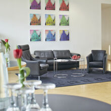 Modernes Wohnzimmer mit schwarzem Ledersofa und -sessel auf einem dunklen Teppich, Glastischen mit Wasserflaschen und Gläsern, einer roten Tulpe in einer Vase und bunten Pop-Art-Gemälden an einer weißen Wand im Hintergrund.