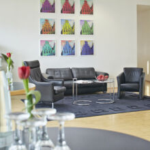 Modernes Wohnzimmer mit schwarzem Ledersofa und -sessel auf einem dunklen Teppich, Glastischen mit Wasserflaschen und Gläsern, einer roten Tulpe in einer Vase und bunten Pop-Art-Gemälden an einer weißen Wand im Hintergrund.