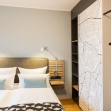 Ein modernes Hotelzimmer mit einem sauber gemachten Doppelbett, zwei an der Wand befestigten Leselampen, einem gemusterten Bettläufer, hellblauen Akzentkissen, offenen Regalen und einem Kleiderschrank mit Kleiderbügeln und einem Stadtplan an der Tür.