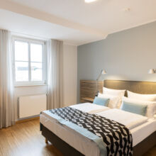 Ein modernes Hotelzimmer mit einem Doppelbett, einer geometrisch gemusterten Decke, zwei Nachttischlampen, hellem Holzfußboden, einem Schreibtisch mit Stuhl und Büchern, einem Fenster mit weißen Vorhängen und neutralen Wänden.