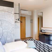 Ein modernes Hotelzimmer mit einem Bett, geometrisch gemustertem Bettzeug, einer Wandkarte, einem Fernseher, einem kleinen Kühlschrank, Wasserflaschen, einer Ablagefläche und einer Holztür. Die Einrichtung ist minimalistisch mit hellen Wänden und Holzakzenten.