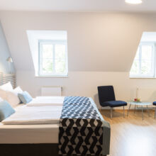 Ein helles, modernes Hotelzimmer mit einem großen Bett, weißer Bettwäsche, blauen und grauen Kissen, zwei blauen Stühlen, einem kleinen runden Tisch mit Obst, Holzfußboden und zwei Fenstern, die Tageslicht hereinlassen.