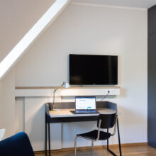 Ein kleiner, moderner Arbeitsbereich mit einem Schreibtisch mit Laptop, Schreibtischlampe und Stuhl unter einem an der Wand montierten Fernseher. In der Nähe befinden sich ein dunkler Kleiderschrank, ein blauer Stuhl und ein Fenster, das Tageslicht hereinlässt.