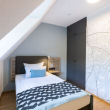 Ein modernes Schlafzimmer mit einem Einzelbett, geometrisch gemusterter Bettwäsche, einer blauen Akzentwand, einem Einbauschrank mit Landkartenmotiv und reichlich natürlichem Licht durch ein Fenster auf der linken Seite.