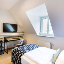 Ein aufgeräumtes, modernes Schlafzimmer mit einem Einzelbett, einer geometrisch gemusterten Decke, einem Schreibtisch mit Stuhl, einem an der Wand montierten Fernseher, einem kleinen Kühlschrank und einem Fenster, das Tageslicht hereinlässt.