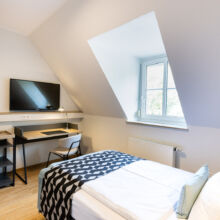 Ein aufgeräumtes, modernes Schlafzimmer mit einem Einzelbett, einer geometrisch gemusterten Decke, einem Schreibtisch mit Stuhl, einem an der Wand montierten Fernseher, einem kleinen Kühlschrank und einem Fenster, das Tageslicht hereinlässt.