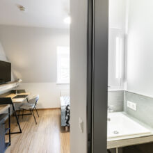 Ein modernes Hotelzimmer mit Schreibtisch, Stuhl, Fernseher, Bett und Fenster; das Badezimmer ist teilweise sichtbar, mit Waschbecken und beleuchtetem Spiegel, durch eine Wand vom Schlafzimmer getrennt.