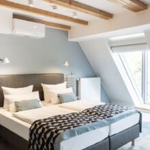 Ein modernes, helles Schlafzimmer mit einem Doppelbett, einer geometrisch gemusterten Decke, zwei an der Wand befestigten Leselampen, Holzbalken an der Decke, hellblauen Wänden und großen Fenstern, die viel Tageslicht hereinlassen.