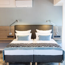 Ein modernes Hotelzimmer verfügt über ein ordentlich gemachtes Doppelbett mit weißer Bettwäsche, grauen und blauen Akzentkissen, Wandlampen und einem Kopfteil aus Holz. Das Zimmer hat eine hellgraue Wand und eine Klimaanlage über dem Bett.