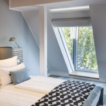 Ein modernes Schlafzimmer mit einem Doppelbett, weißer Bettwäsche, einer geometrisch gemusterten Decke und grauen Akzent-Kissen. Es gibt ein Fenster mit Blick auf grüne Bäume und eine Wandlampe neben dem Bett.