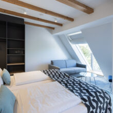 Modernes Hotelzimmer mit einem Doppelbett, einer geometrisch gemusterten Decke, einem grauen Sofa, einem kleinen runden Glastisch, einem dunklen Stuhl, einem offenen Regal mit einem Safe und einem großen Fenster mit schräger Decke und Holzbalken.