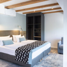 Modernes Hotelzimmer mit einem Doppelbett, einer geometrisch gemusterten Decke, zwei Nachttischlampen, einem offenen Kleiderschrank, einem grauen Sofa und einem runden Glastisch. Das Zimmer hat hölzerne Deckenbalken und eine weiche, neutrale Farbgebung.