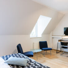 Ein helles, modernes Zimmer mit zwei blauen Stühlen und einem runden Glastisch am Fenster, einem Schreibtisch mit einem Stuhl und einem montierten Fernseher sowie einem Teil eines Bettes mit einer geometrisch gemusterten Decke und einem Kissen im Vordergrund.