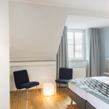 Ein modernes Hotelzimmer mit einem Doppelbett, zwei blauen Stühlen, einem Fernseher auf einem Schrank, hellem Holzfußboden, einer kleinen Würfellampe und einem Fenster mit grauen Vorhängen, durch das Tageslicht fällt.