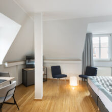 Ein modernes, helles Hotelzimmer mit einem Doppelbett, einem Schreibtisch und einem Stuhl, zwei blauen Stühlen, einem kleinen Fernseher auf einem Schrank, Holzfußboden und großen Fenstern mit grauen Vorhängen, die natürliches Licht hereinlassen.